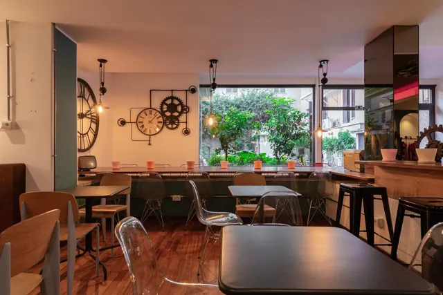 A dining room with wooden and clear chairs at tables, a large wall clock with gears, and a window looking onto a courtyard with trees and plants.