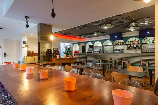 A bar area with a long wooden counter in the foreground, arched shelves of bottles, two wall-mounted televisions, and tables and chairs.