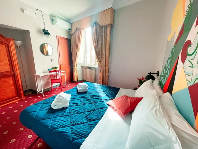 A bedroom with a teal quilted bedspread, white pillows, a red cushion, a colourful painted headboard wall, a white desk with a red chair, and a wooden wardrobe near a window with curtains.