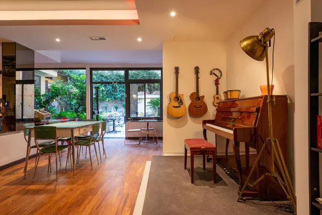 A room with a green dining table and green chairs, three guitars and an upright piano against the walls, and glass doors looking out onto a garden with trees.