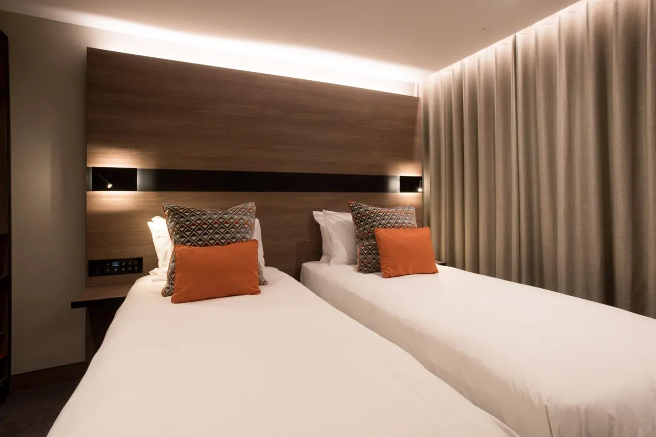 Two single beds with white linen and orange and patterned cushions sit in front of a wood-panelled headboard wall, with floor-to-ceiling curtains on the right.