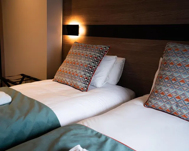 Two single beds with white linen, patterned cushions and a dark wooden headboard lit by a wall light, in a bedroom.