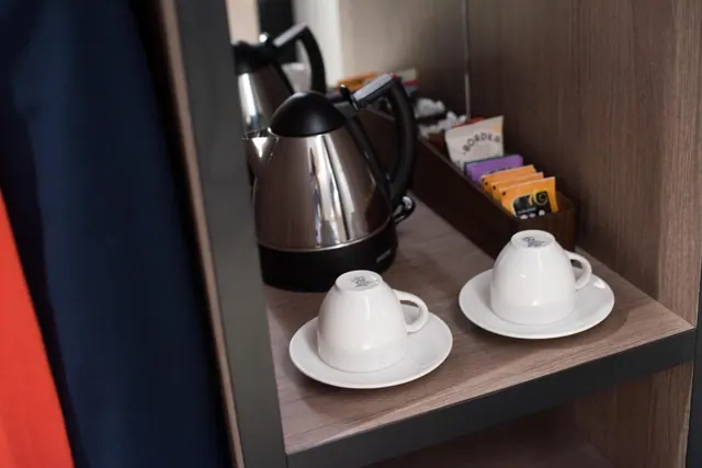 Two stainless steel kettles, two white cups on saucers and a holder of tea sachets sit on a wooden shelf.