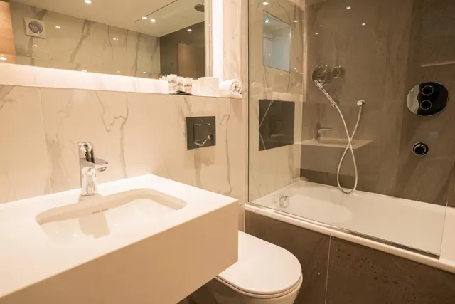 A modern bathroom with marble-look wall tiles, a single wash basin, a wall-mounted toilet and a bathtub with a handheld shower behind a glass screen.