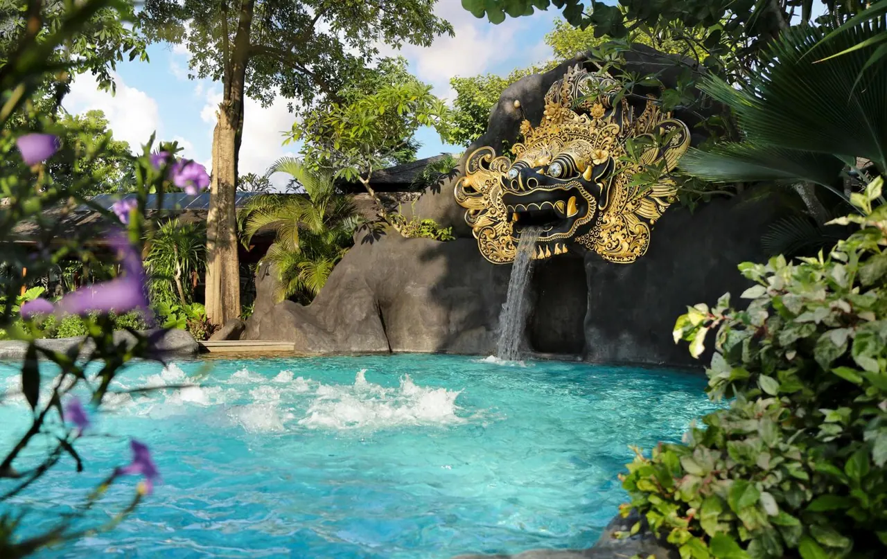 A turquoise pool with a waterfall spilling from a gilded Barong mask sculpture on a dark rock wall, framed by tropical plants and trees.
