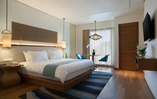 A bedroom with a king bed, a pair of blue chairs by a window with sheer curtains, a wall-mounted television and a wooden door.