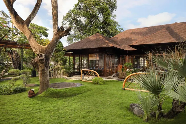 A trimmed lawn garden with a large bare-branched tree, bamboo benches, palms, and a brown-roofed building with glass walls under a partly cloudy sky.