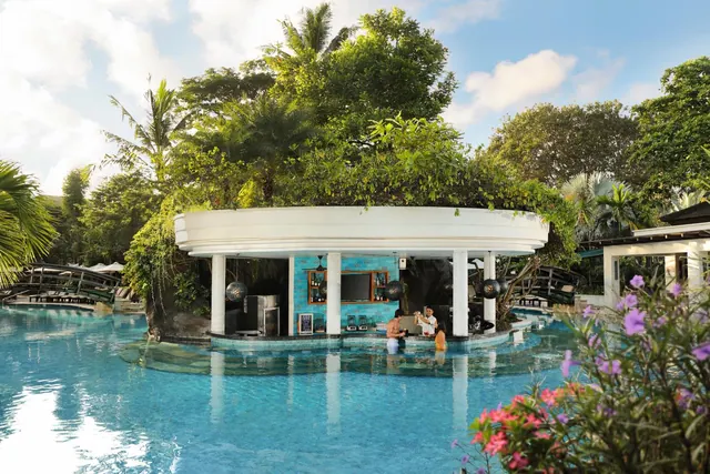 A swim-up bar with a white rounded roof and turquoise wall sits in a large pool surrounded by tropical trees, with a few people standing in the water.