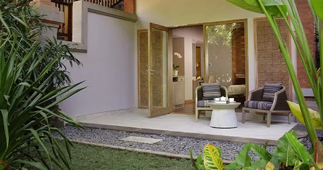 A paved terrace outside open wooden doors has two wicker armchairs with striped cushions, a round white side table, and a gravel border planted with tropical foliage.