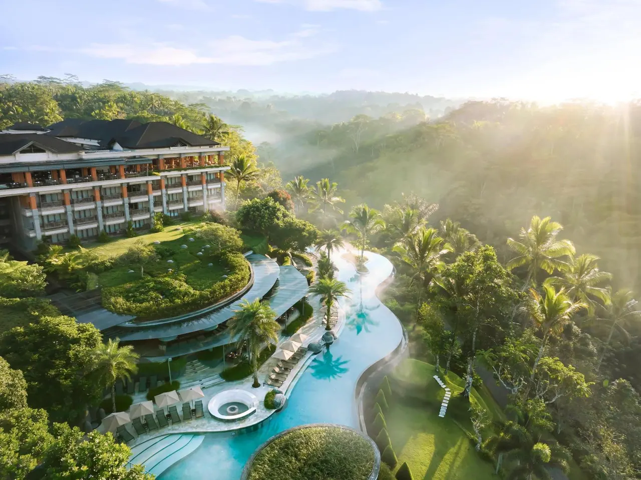 Aerial view of a resort with a winding pool, sun loungers under white umbrellas, a multi-storey building, and misty jungle hills beyond.