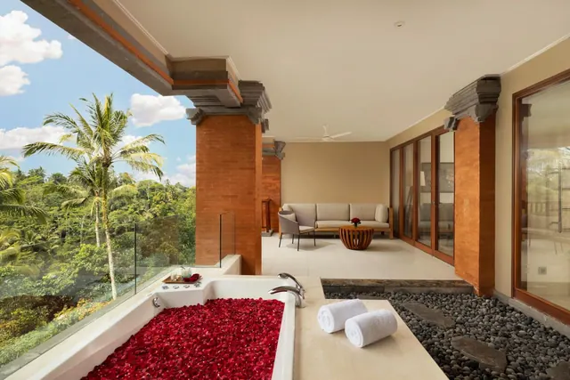 An open-air bathtub filled with red rose petals sits beside a glass railing overlooking palm trees and jungle, with a seating area and brick pillars nearby.