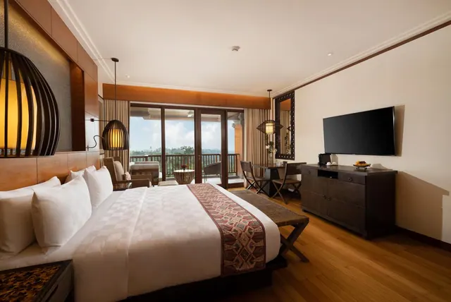 A bedroom with a king bed, a wall-mounted television on a wooden dresser, and sliding glass doors opening onto a balcony with a view of trees.