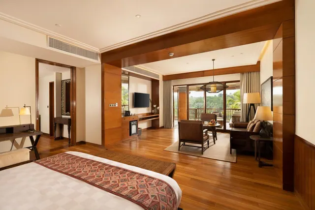 A spacious bedroom with a large bed in the foreground, a seating area with armchairs and a sofa, and sliding glass doors opening onto a balcony with trees beyond.