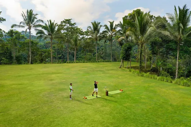 Aerial view of a grassy golf driving range where two men are hitting from mats, bordered by palm trees and dense jungle.