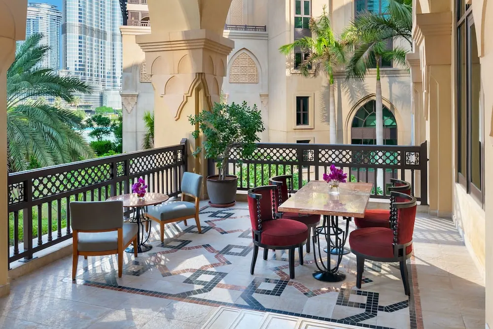A covered terrace with arched columns, a patterned tiled floor, red upholstered chairs and tables, with palm trees and skyscrapers visible beyond the lattice railing.