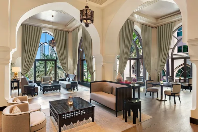 A lounge area with beige sofas, armchairs and low carved coffee tables under arched ceilings, with tall windows showing palm trees and high-rise towers outside.