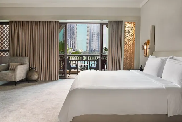 A bedroom with a made king bed, an armchair, and a balcony door showing high-rise towers outside.