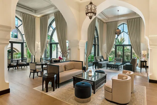 A lounge area with arched openings, grey floor-length curtains, upholstered sofas and armchairs, a hanging lantern, and palm trees visible through tall windows.
