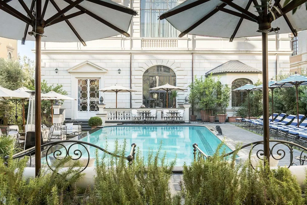 A courtyard swimming pool with blue-cushioned loungers and white umbrellas, with a pale stone palazzo-style building behind it.