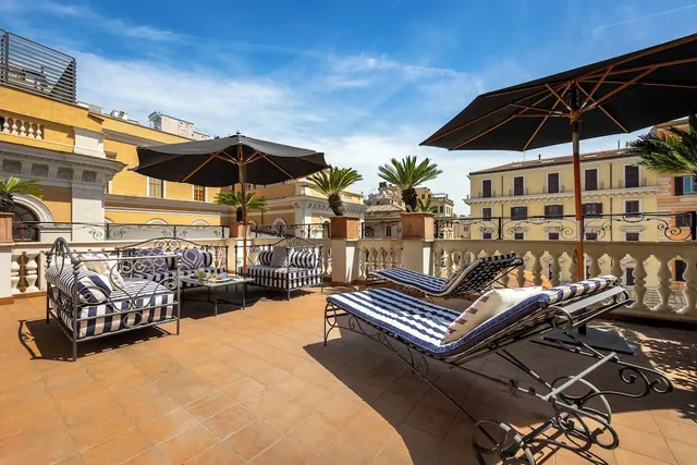 A rooftop terrace with striped wrought-iron sofas and sun loungers, shaded by dark parasols, with palm trees and yellow apartment buildings beyond the balustrade.