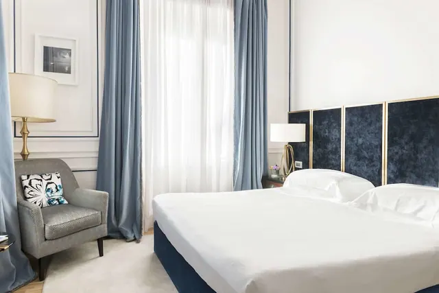 A bedroom with a made double bed, a dark blue velvet headboard, blue curtains and a grey armchair beside a window.