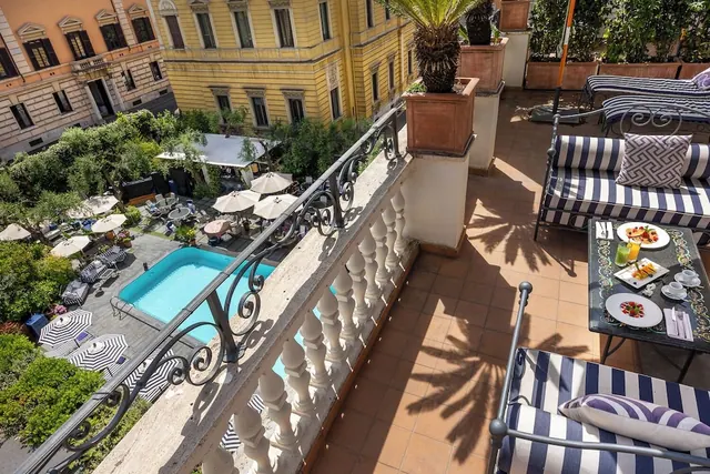 A terrace with striped lounge seating and a set table overlooks a hotel pool, umbrellas and garden below.