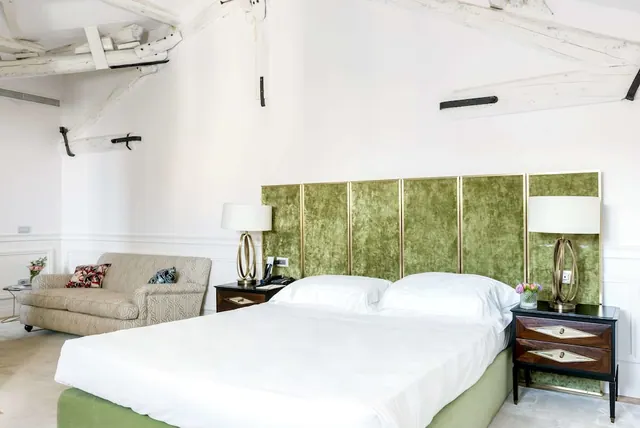 A bedroom with a made double bed, green velvet headboard panels, two bedside lamps and a patterned sofa beneath exposed white ceiling beams.