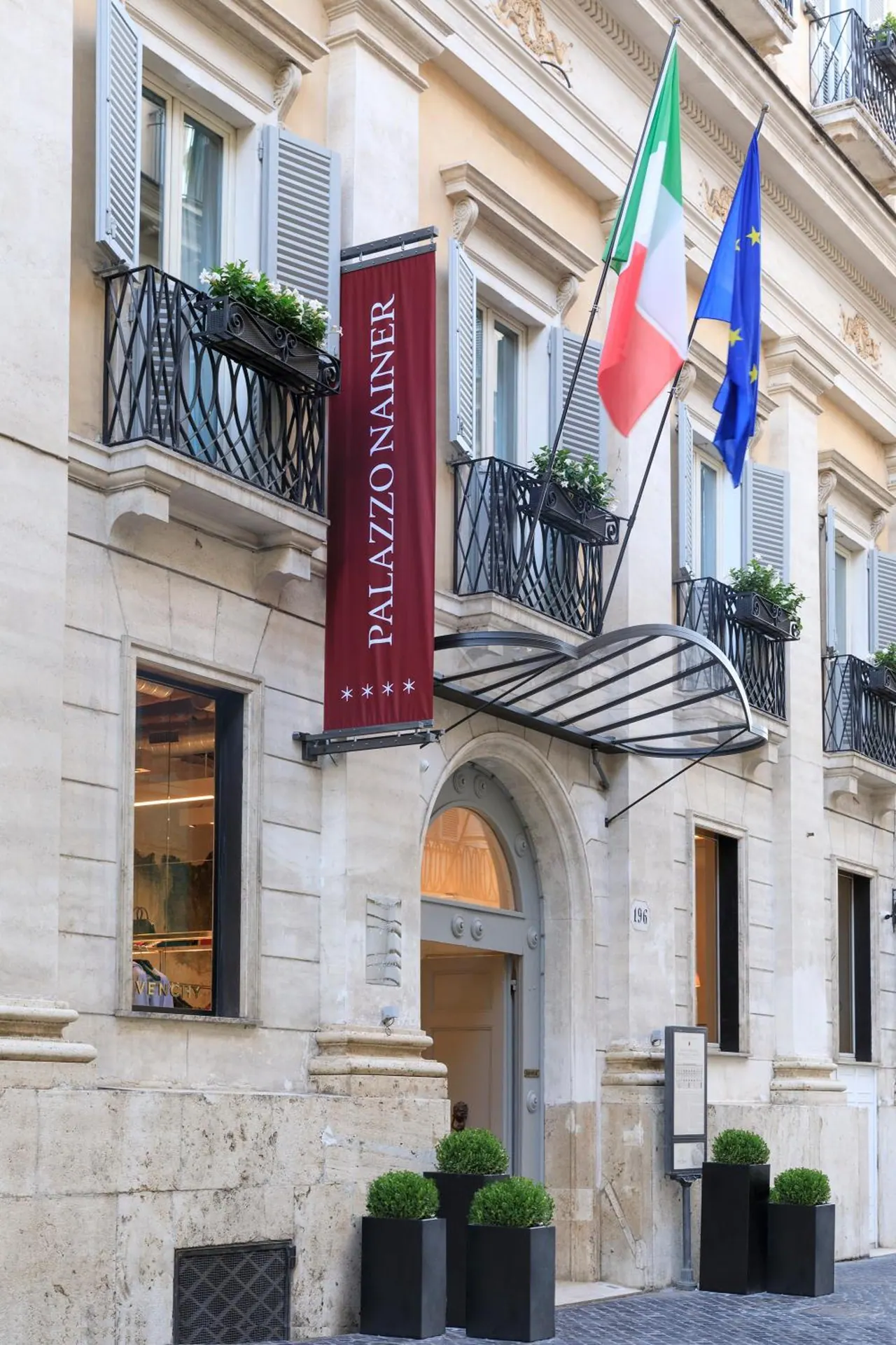 Street-level facade of Palazzo Nainer with a red vertical banner, Italian and EU flags, an arched entrance, and boxwood planters on the pavement.