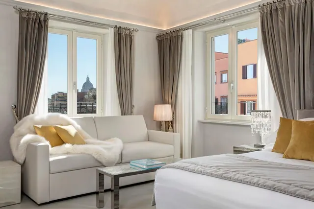 A bedroom with a made bed, a white sofa with a fur throw and gold cushions, and two windows looking out over rooftops to a domed basilica.