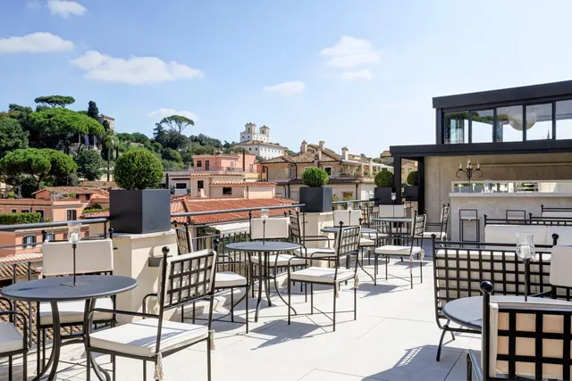 A rooftop terrace with black metal tables and cushioned chairs overlooks terracotta rooftops, pine trees and a hillside under a blue sky.