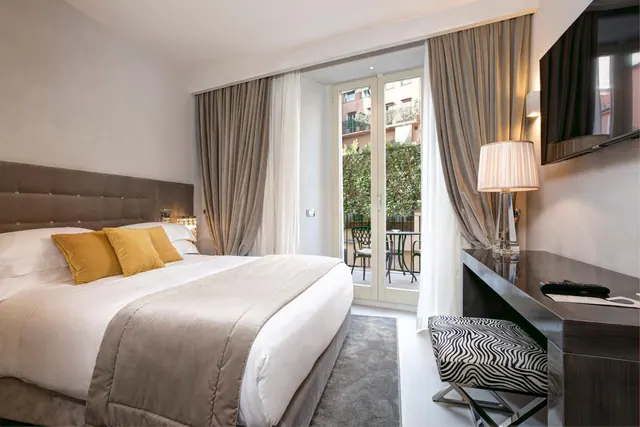 A bedroom with a large bed, yellow cushions, a wall-mounted television, a desk, and open French doors onto a balcony with a bistro table and chairs.