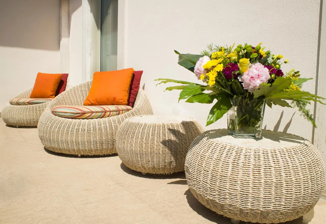 A sunlit terrace with two woven wicker armchairs with orange cushions, round wicker ottomans and a vase of flowers on a low table.