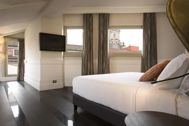 A bedroom with a made white bed, a wall-mounted flat-screen television, and windows looking out over rooftops and a domed church.