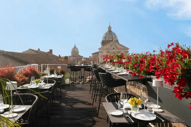 A rooftop terrace restaurant with set tables and red flowering plants in planters, with church domes and rooftops visible in the background.