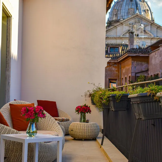 A terrace with wicker armchairs, red cushions, a vase of pink flowers and planters along the railing, with the dome of St. Peter's Basilica visible above the neighbouring rooftops.