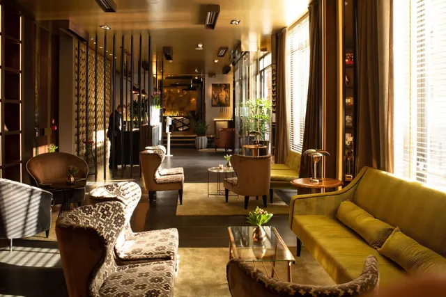 A lounge area with upholstered armchairs, a yellow velvet sofa, a glass coffee table and sunlight through window blinds, with a staff member visible at a reception desk in the background.