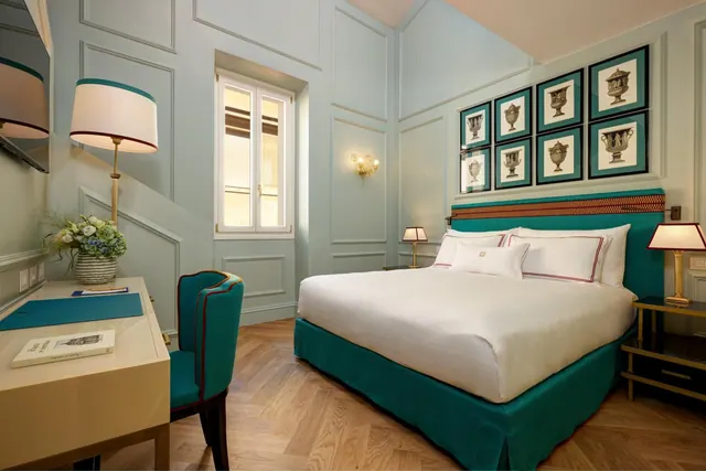 A bedroom with a king bed with a teal base and headboard, framed urn prints on the wall, a writing desk with a teal chair, and a window on the left.