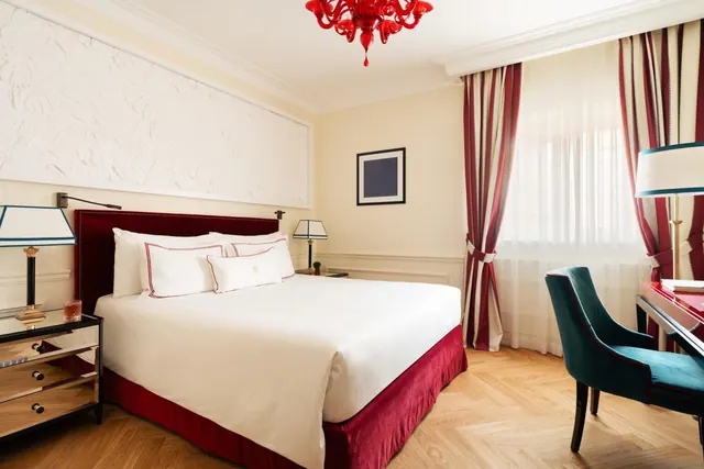 A bedroom with a king bed, red velvet headboard, red glass chandelier, striped curtains and a teal chair at a desk by the window.