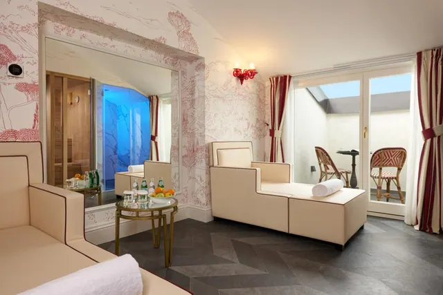 A staged lounge room with cream sofas, red toile wallpaper, a glass table with fruit and water bottles, a sauna visible in a mirror, and a terrace with bistro chairs beyond the glass doors.