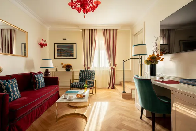 A living area in a hotel suite with a red velvet sofa, a patterned armchair, a coffee table with croissants and coffee, a desk with a teal chair, and a wall-mounted television.
