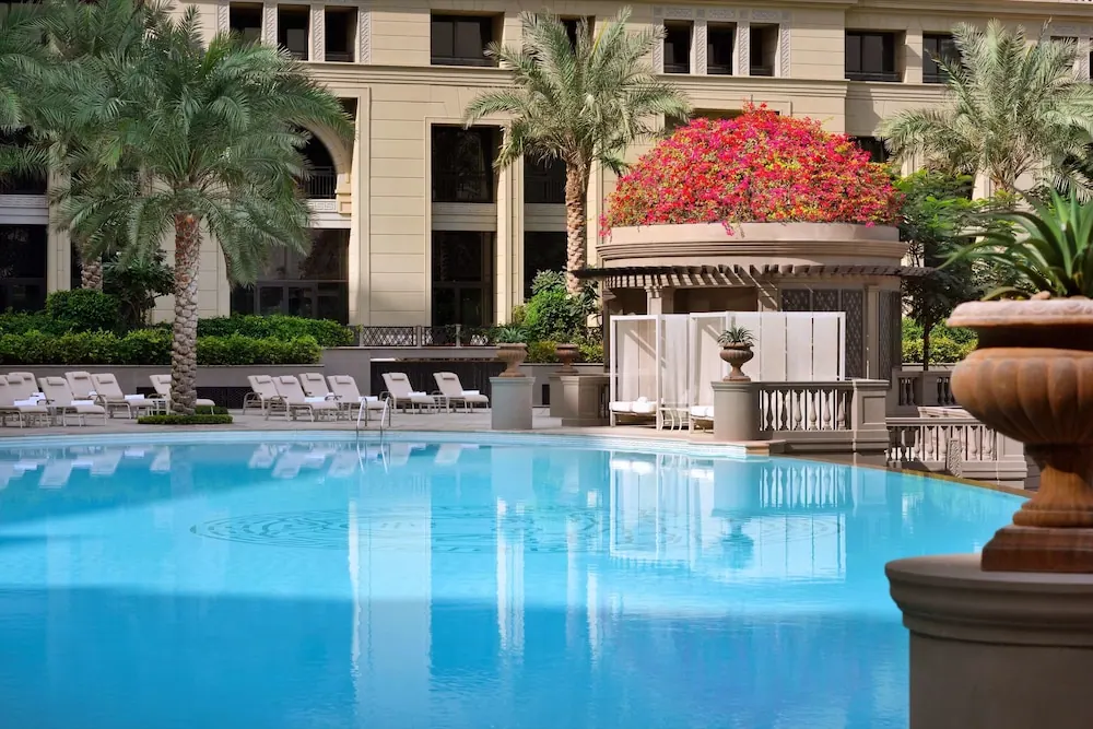A large swimming pool with white sun loungers, palm trees and a flowering planter in front of a cream-coloured building.