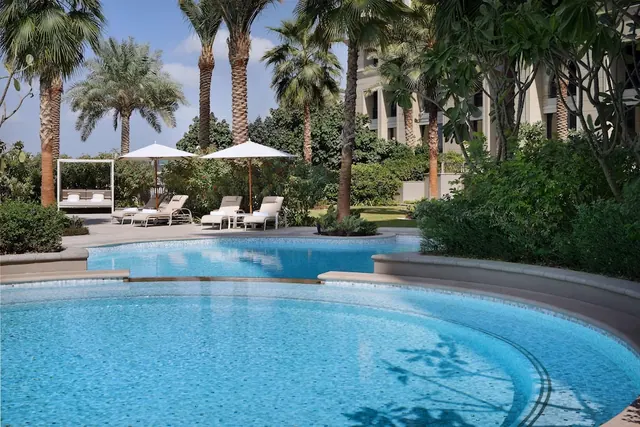 A curved outdoor pool with sun loungers and white umbrellas on the deck, surrounded by palm trees and green shrubs, with a building on the right.