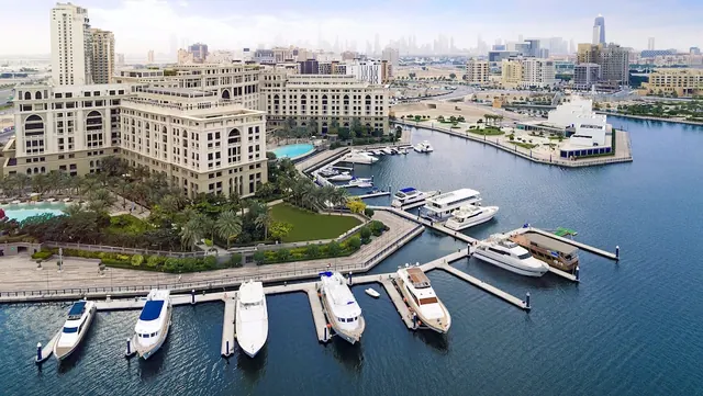 Aerial view of a cream-coloured hotel building beside a marina with moored yachts and a city skyline in the background.
