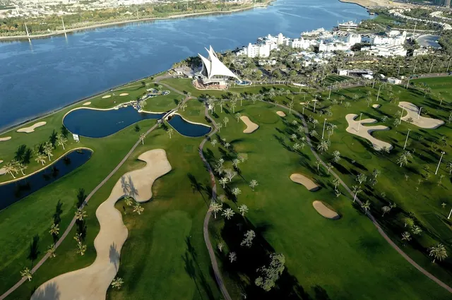 Aerial view of a golf course with sand bunkers, ornamental ponds and a white sail-shaped building beside a waterway.