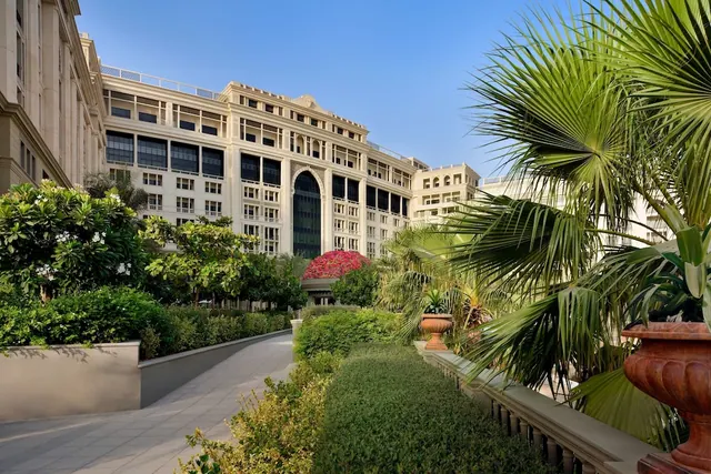 A paved walkway runs between clipped hedges, palm plants and stone urns toward a large cream-coloured building with many windows under a clear blue sky.