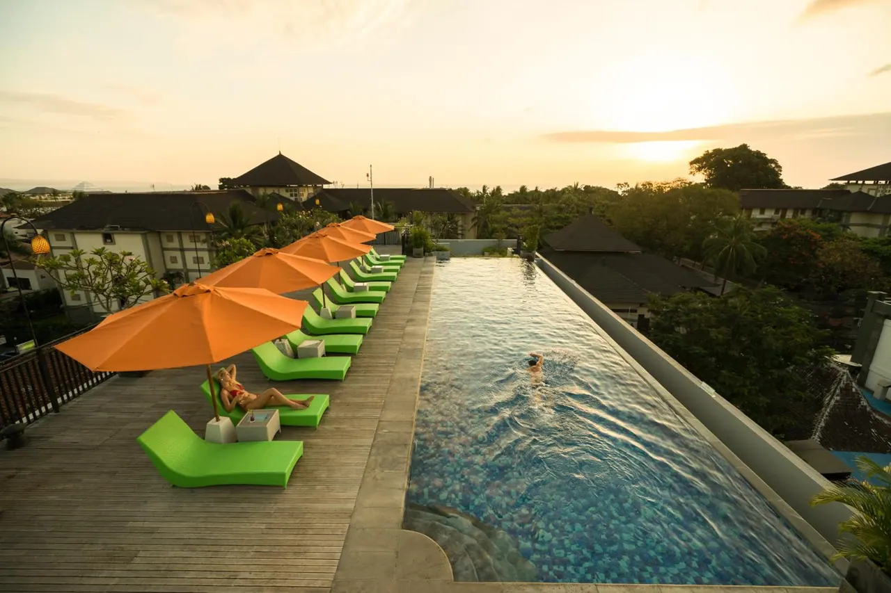 Rooftop infinity pool at sunset with green loungers and orange umbrellas, a woman lying on a lounger and a swimmer in the water.
