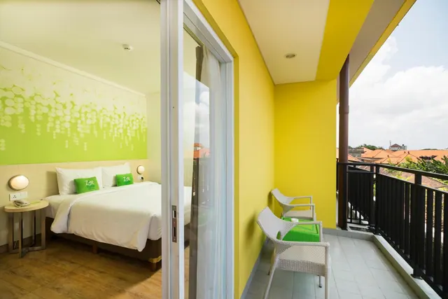 A balcony with two white wicker chairs and a green side table, with a double bed in the room visible through the sliding glass door and rooftops beyond.