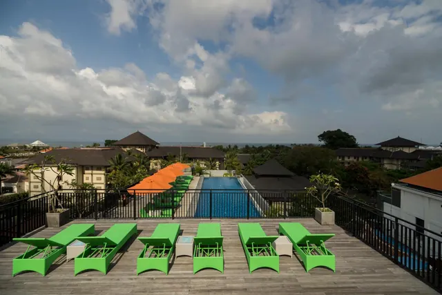 A rooftop deck with green sun loungers and orange umbrellas overlooks a swimming pool, surrounding rooftops and a strip of sea on the horizon.