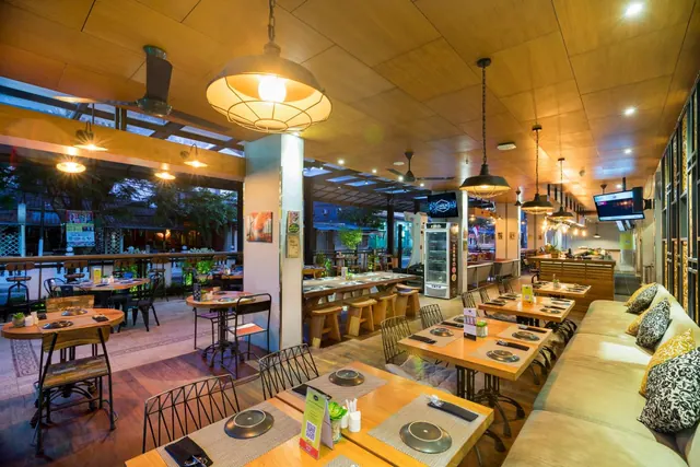 A restaurant dining room with wooden tables set with plates, pendant lamps, a sofa bench along the right wall, and trees visible through the open side.
