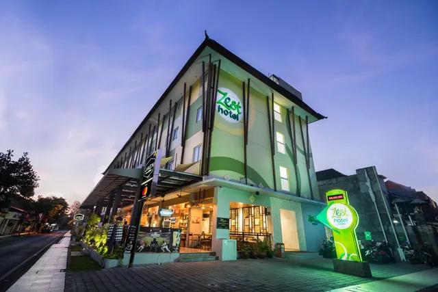 The exterior of Zest Hotel at dusk, with its green facade, illuminated signs and motorbikes parked along the street.